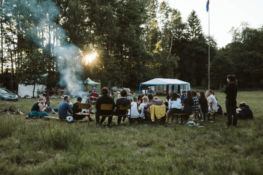 A group of adults relaxing around a campfire in a scenic outdoor setting with tents.